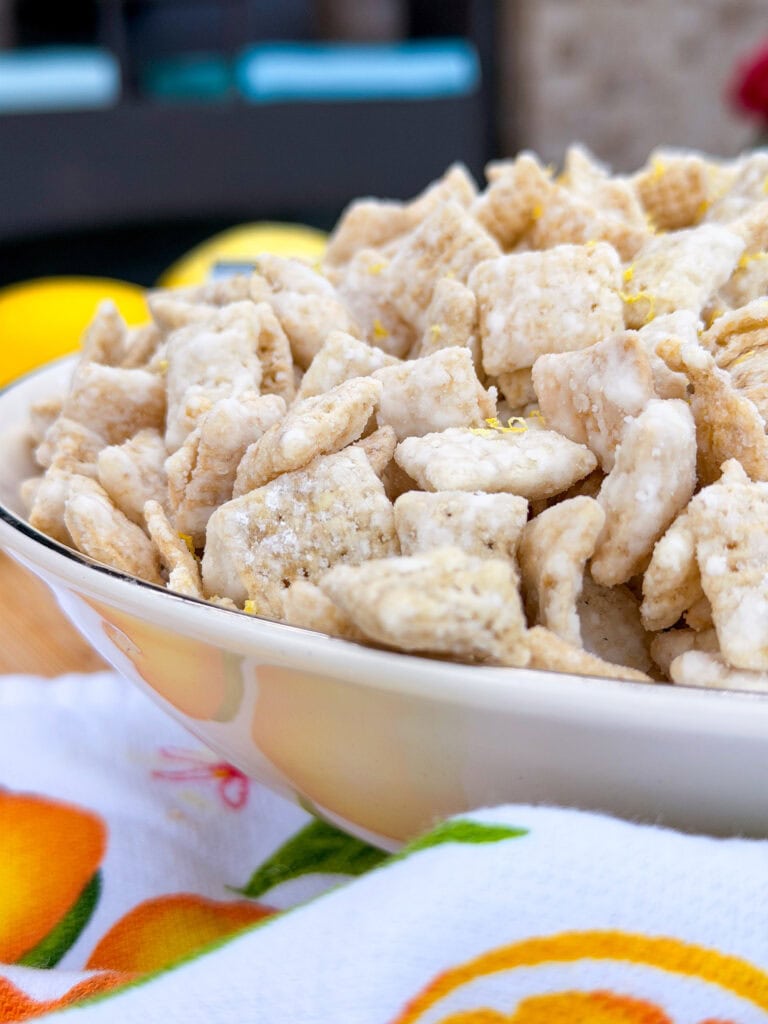 A close-up of a white bowl filled with powdered cereal pieces, sitting on a colorful cloth with fruit designs. The background is blurred.