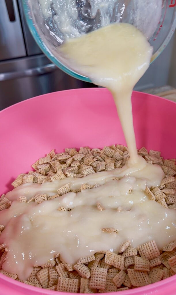 A bowl of Chex cereal in a pink bowl has creamy melted mixture being poured over it from a glass bowl.