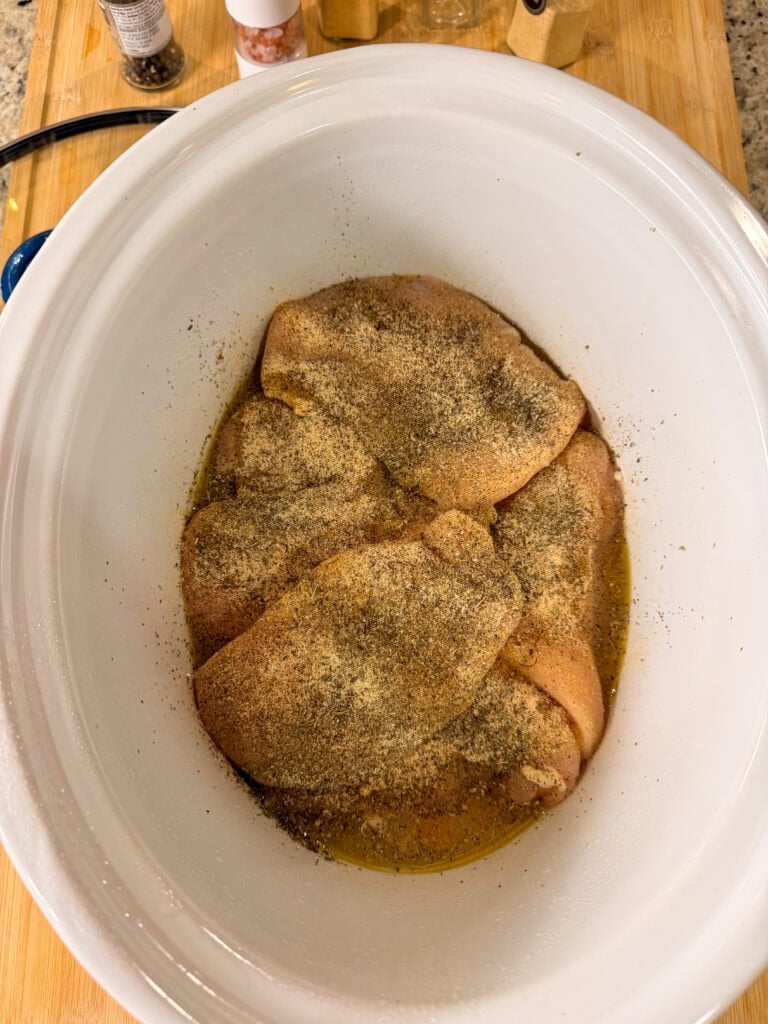 Raw chicken breasts in a white slow cooker, seasoned with various spices and sitting in a small amount of liquid on a wooden countertop.