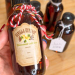 A hand holds a small bottle labeled Vanilla Extract Homemade with love. Several similar bottles with tags and twine are on a wooden surface in the background.
