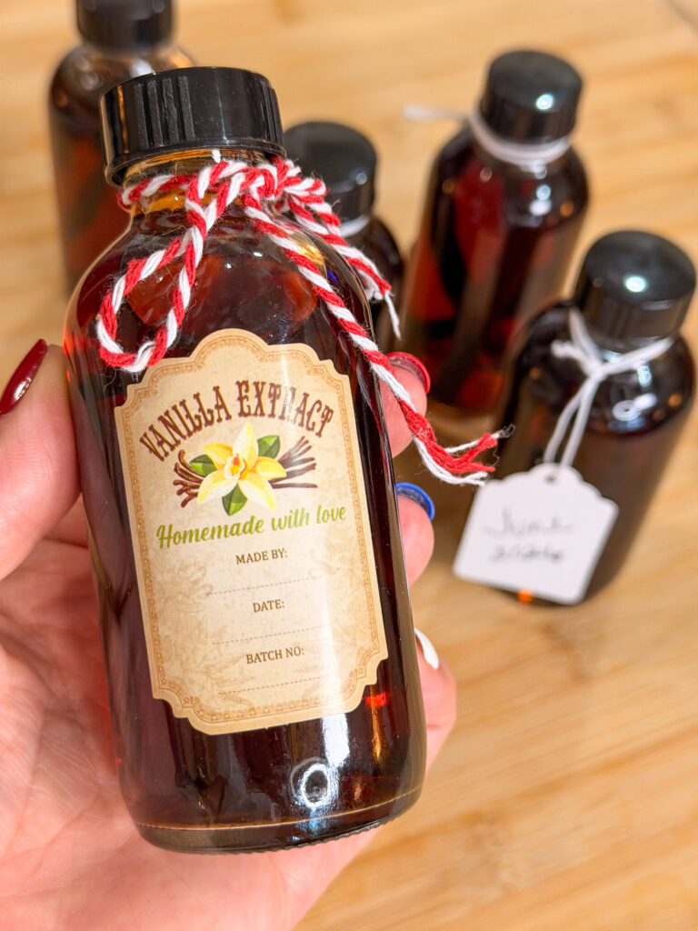 A hand holds a small bottle labeled Vanilla Extract Homemade with love. Several similar bottles with tags and twine are on a wooden surface in the background.