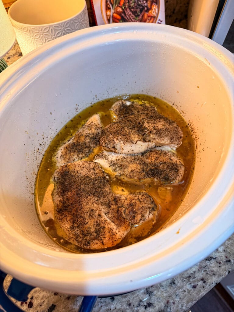 Four seasoned chicken breasts cooking in a white slow cooker with broth visible at the bottom. The slow cooker sits on a kitchen counter with various items in the background.