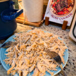 A plate of shredded chicken sits on a granite countertop beside two forks, a slow cooker, a white textured canister, and a cookbook titled The Stress-Free Slow Cooker in the background.