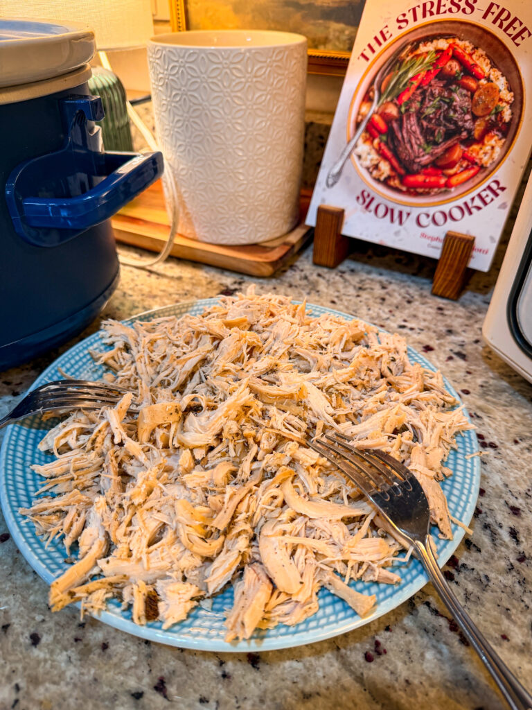 A plate of shredded chicken sits on a granite countertop beside two forks, a slow cooker, a white textured canister, and a cookbook titled The Stress-Free Slow Cooker in the background.