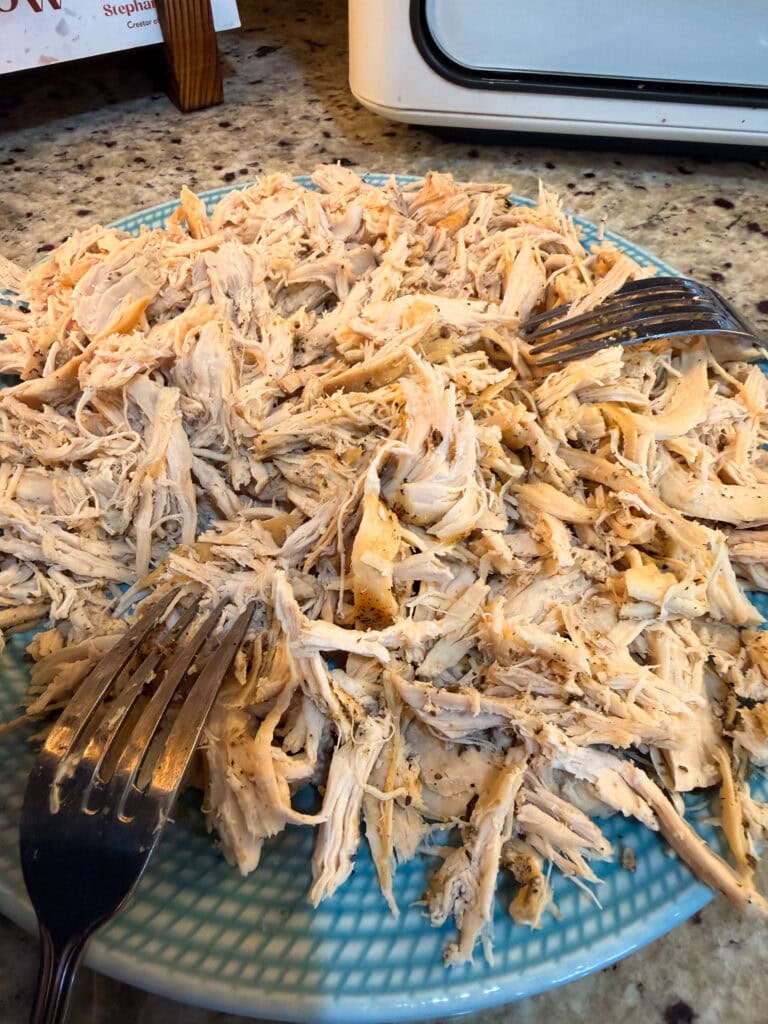 A blue plate piled with shredded, seasoned chicken sits on a kitchen counter. Two metal forks rest in the chicken, used for pulling it apart. A toaster and recipe book are visible in the background.
