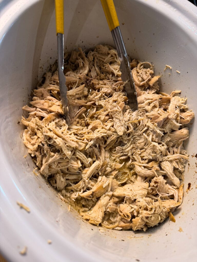 Shredded cooked chicken in a white slow cooker with metal tongs and some broth at the bottom.