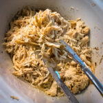 Shredded chicken being lifted with metal tongs from a white slow cooker.