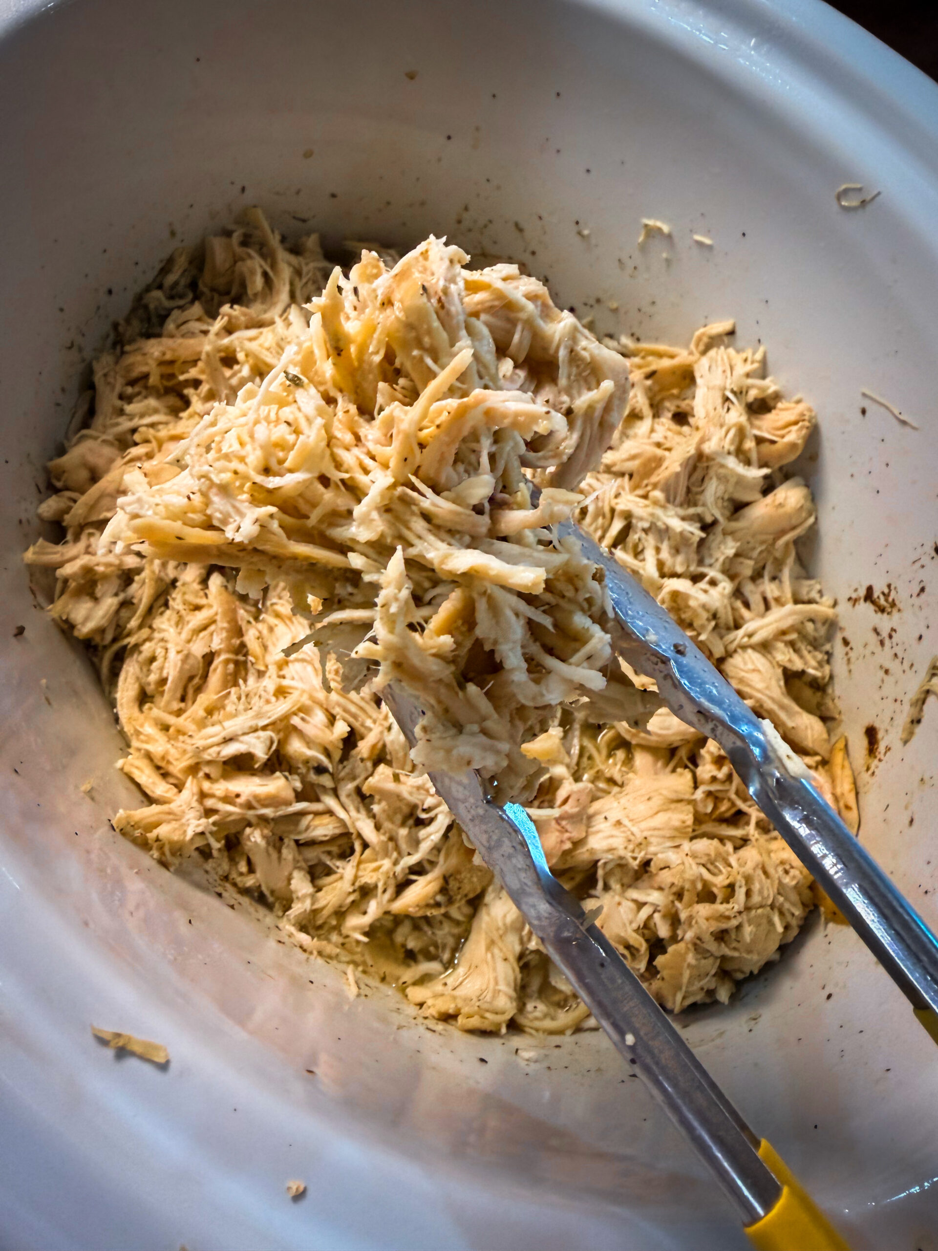 Shredded chicken being lifted with metal tongs from a white slow cooker.