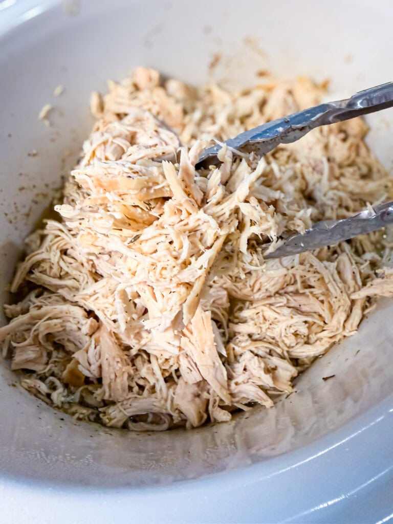 Shredded chicken being lifted with metal tongs from a white slow cooker, with the chicken appearing moist and tender.