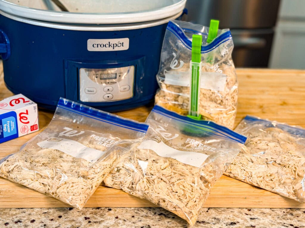Four freezer bags filled with shredded chicken sit on a counter next to a blue Crock-Pot slow cooker and some measuring cups in a kitchen setting.