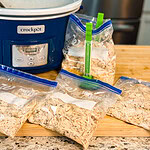 Four freezer bags filled with shredded chicken sit on a counter next to a blue Crock-Pot slow cooker and some measuring cups in a kitchen setting.