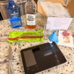 A kitchen counter with a digital scale, a knife, bottles of vodka and bourbon, sugar, a small empty bottle, a blue funnel, a packet, and an open box with bubble wrap.
