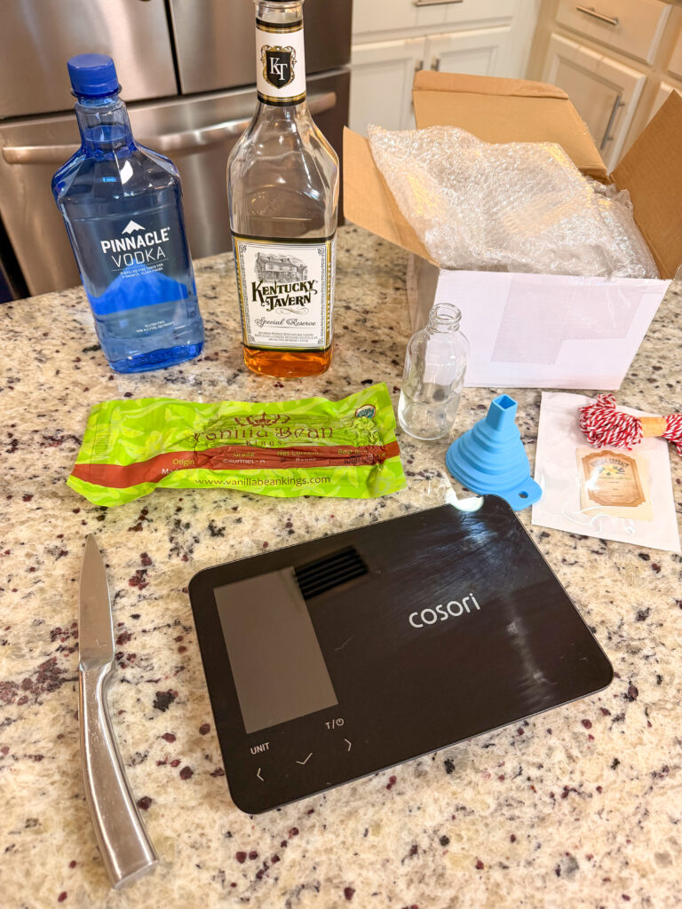 A kitchen counter with a digital scale, a knife, bottles of vodka and bourbon, sugar, a small empty bottle, a blue funnel, a packet, and an open box with bubble wrap.