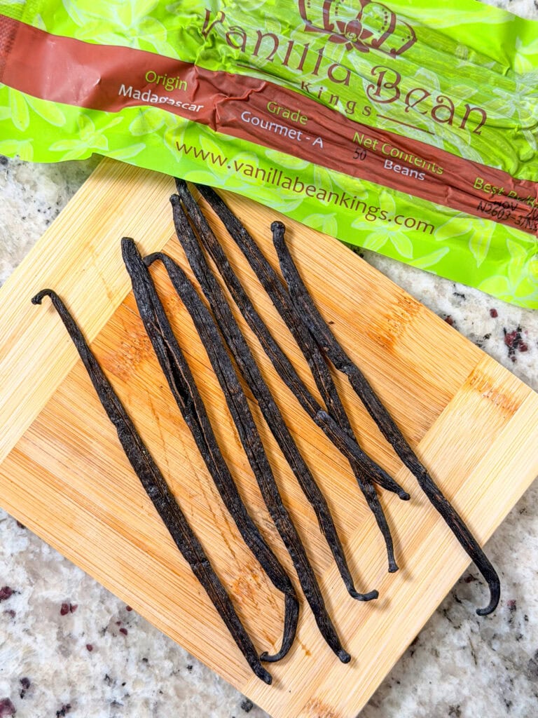 Several dried vanilla beans are arranged on a small wooden cutting board, with a green Vanilla Bean Kings package partially visible in the background. The surface underneath appears to be a granite countertop.