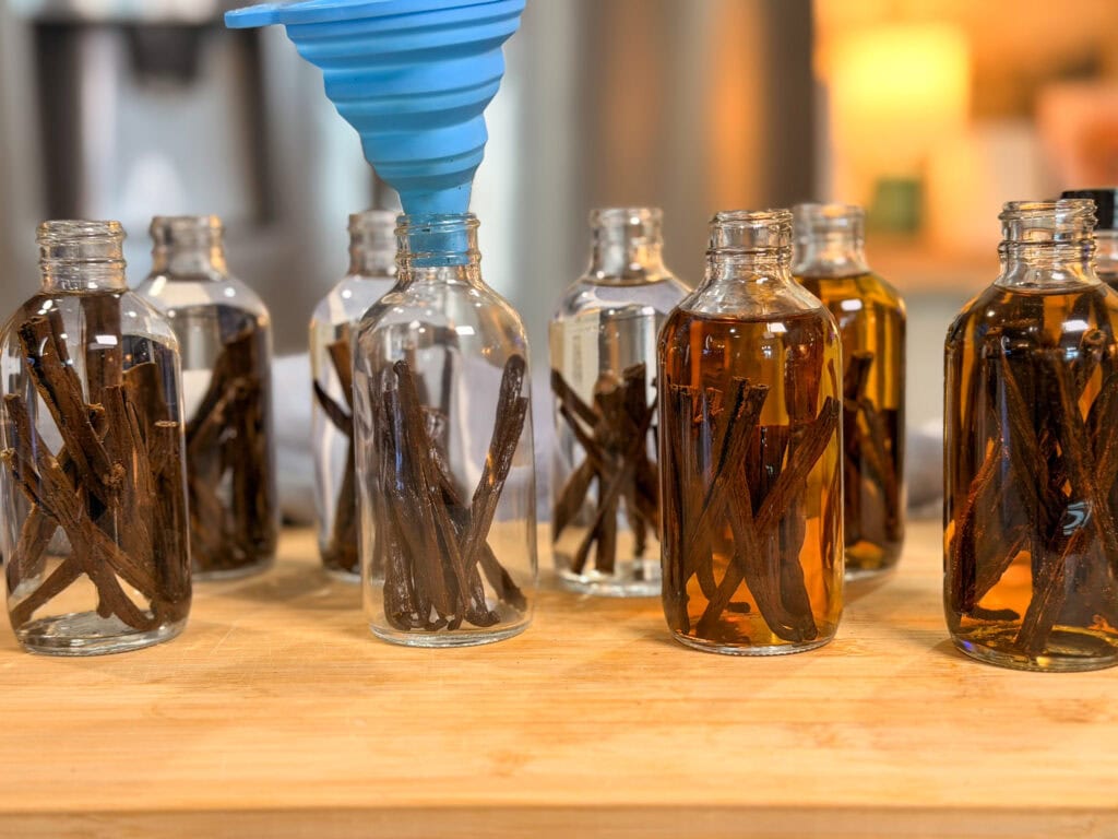 Several glass bottles filled with vanilla beans sit on a wooden surface. Some bottles are being filled with liquid using a blue funnel. Background appears to be a warmly lit kitchen.