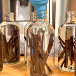 Several glass bottles filled with vanilla beans are arranged on a wooden surface. A blue funnel is positioned above one bottle, as if ready to pour liquid inside. A refrigerator is visible in the blurred background.
