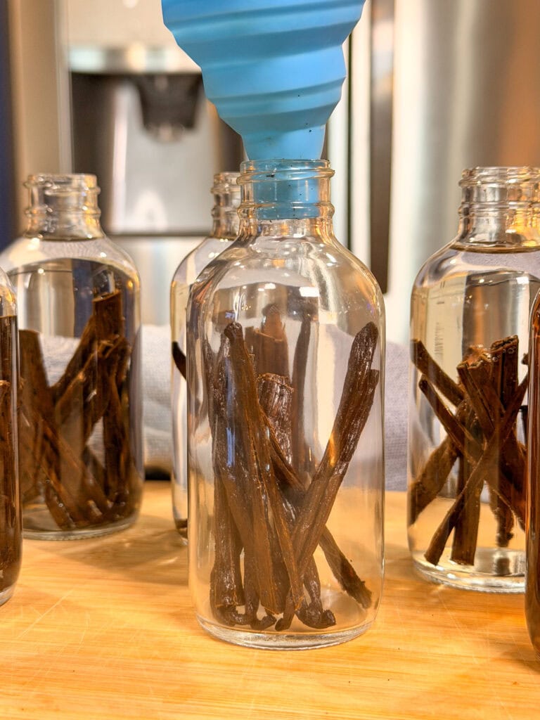 Several glass bottles filled with vanilla beans are arranged on a wooden surface. A blue funnel is positioned above one bottle, as if ready to pour liquid inside. A refrigerator is visible in the blurred background.