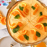 An orange cream pie with a graham cracker crust, topped with orange slices and mint leaves, sliced into eight pieces. Nearby are plates, forks, and a glass of orange juice.