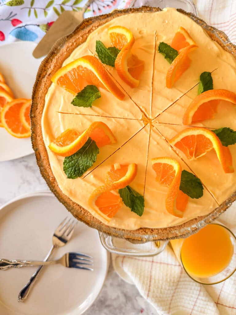 An orange cream pie with a graham cracker crust, topped with orange slices and mint leaves, sliced into eight pieces. Nearby are plates, forks, and a glass of orange juice.