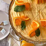 A creamy orange pie garnished with fresh orange slices and mint leaves sits on a table, partially sliced. Plates with forks and more orange slices are nearby.