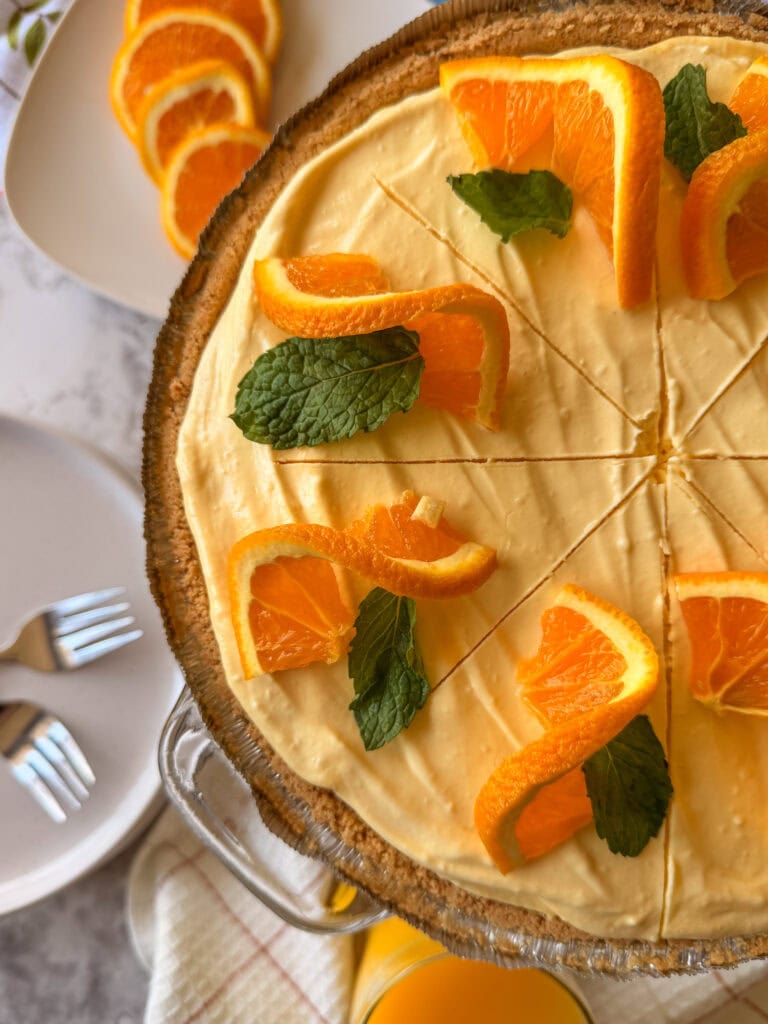 A creamy orange pie garnished with fresh orange slices and mint leaves sits on a table, partially sliced. Plates with forks and more orange slices are nearby.