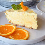 A slice of creamy orange pie with a graham cracker crust is on a plate, garnished with fresh orange slices and a mint leaf. Two more orange slices are on the plate beside the pie.