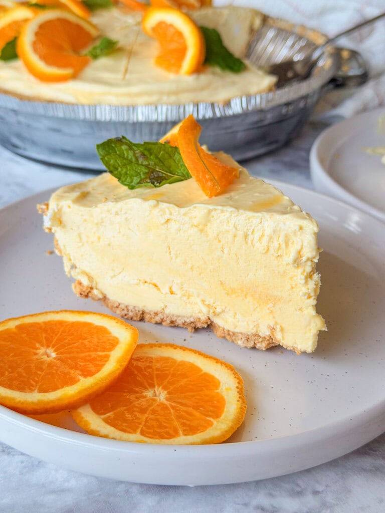 A slice of creamy orange pie with a graham cracker crust is on a plate, garnished with fresh orange slices and a mint leaf. Two more orange slices are on the plate beside the pie.