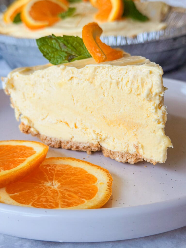 A slice of creamy orange cheesecake with a crumbly crust is served on a white plate, garnished with orange slices and mint leaves. The rest of the cheesecake is in the background.