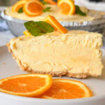 A slice of creamy orange pie with a crumbly crust, garnished with fresh orange slices and mint, sits on a white plate with two additional orange slices in front.