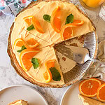 A creamy orange pie with slices cut, topped with fresh orange segments and mint leaves. Two plated slices sit nearby, one with extra orange slices. A glass of orange juice is visible in the corner.