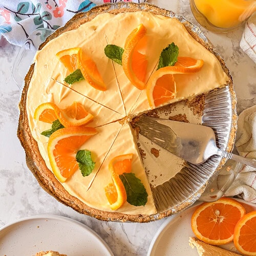 A creamy orange pie with slices cut, topped with fresh orange segments and mint leaves. Two plated slices sit nearby, one with extra orange slices. A glass of orange juice is visible in the corner.