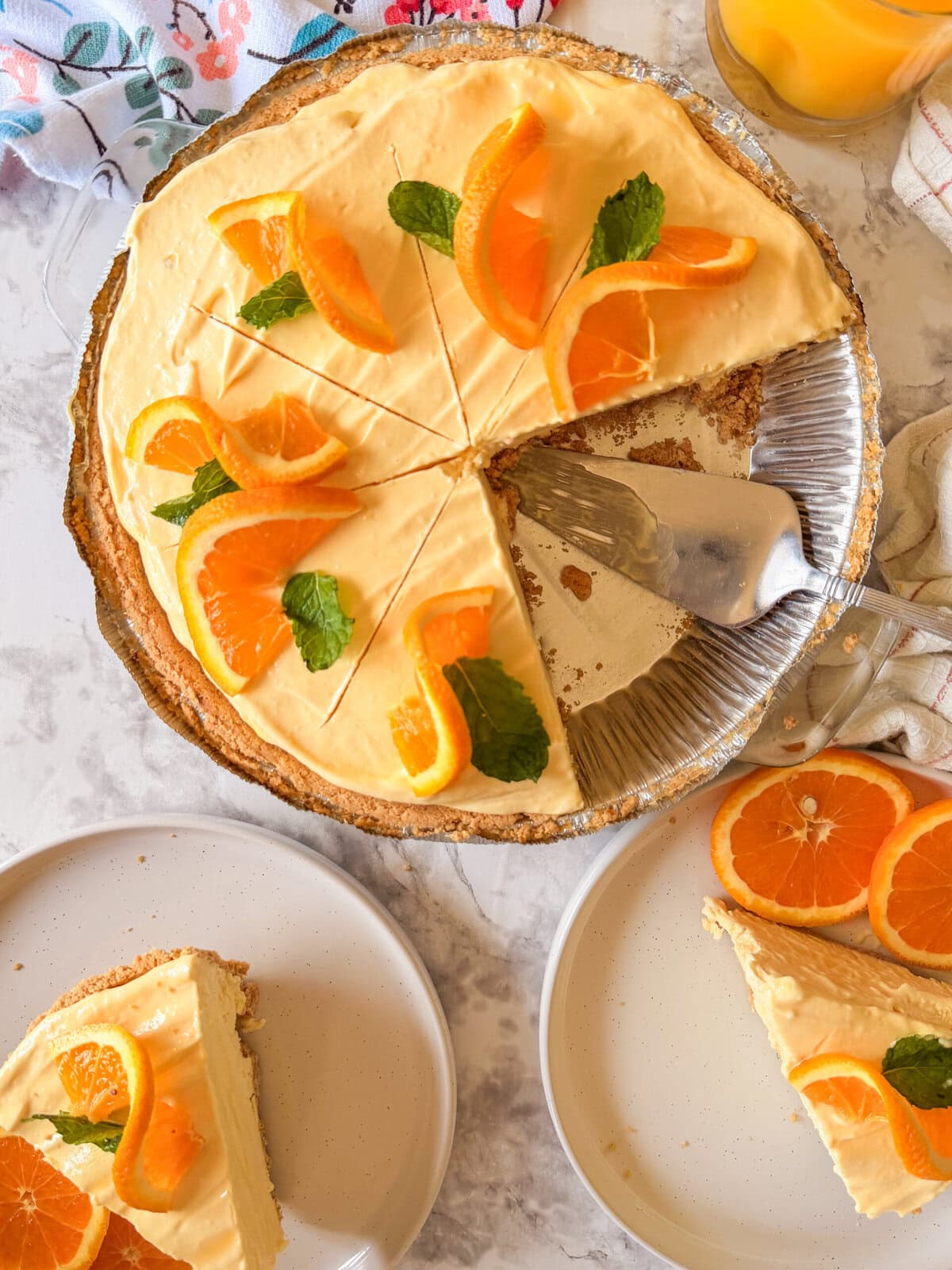 A creamy orange pie with slices cut, topped with fresh orange segments and mint leaves. Two plated slices sit nearby, one with extra orange slices. A glass of orange juice is visible in the corner.