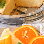 A creamy orange cheesecake with a sliced piece, garnished with fresh orange slices and mint leaves, sits on a glass pie dish. A cake server rests nearby and additional orange slices are in the foreground.