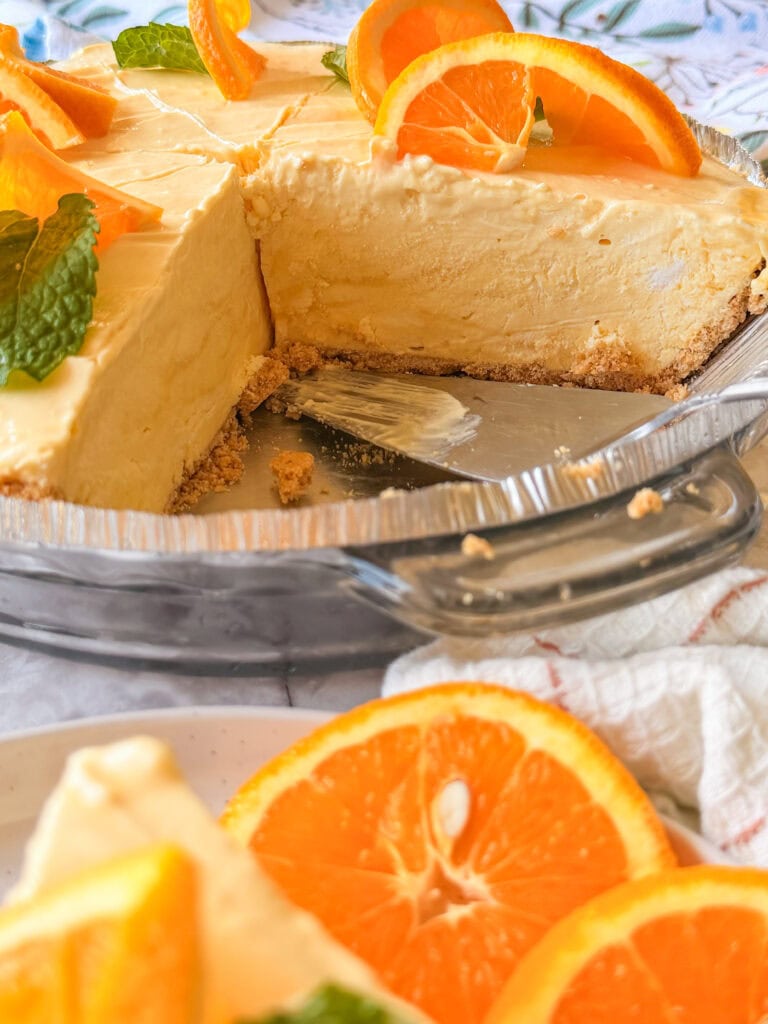 A creamy orange cheesecake with a sliced piece, garnished with fresh orange slices and mint leaves, sits on a glass pie dish. A cake server rests nearby and additional orange slices are in the foreground.