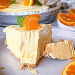 A slice of creamy orange cheesecake with a graham cracker crust, topped with an orange slice and mint, sits on a plate with a forkful removed. More cheesecake and orange slices are in the background.