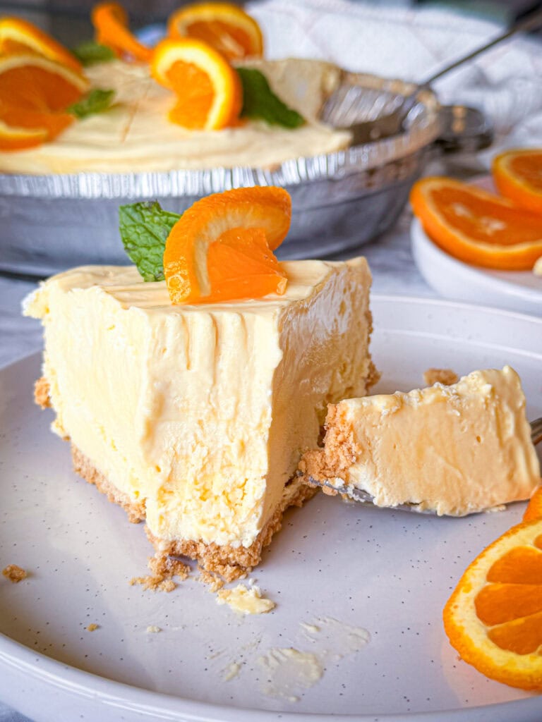 A slice of creamy orange cheesecake with a graham cracker crust, topped with an orange slice and mint, sits on a plate with a forkful removed. More cheesecake and orange slices are in the background.