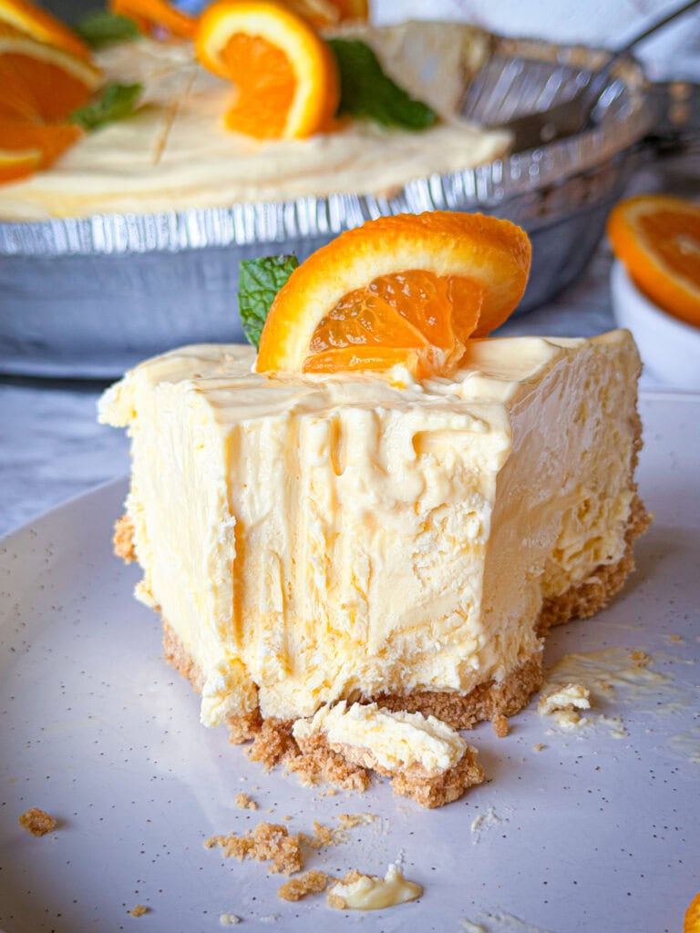 A creamy orange cheesecake slice with a graham cracker crust is topped with an orange slice and mint, placed on a white plate with a bite taken out.
