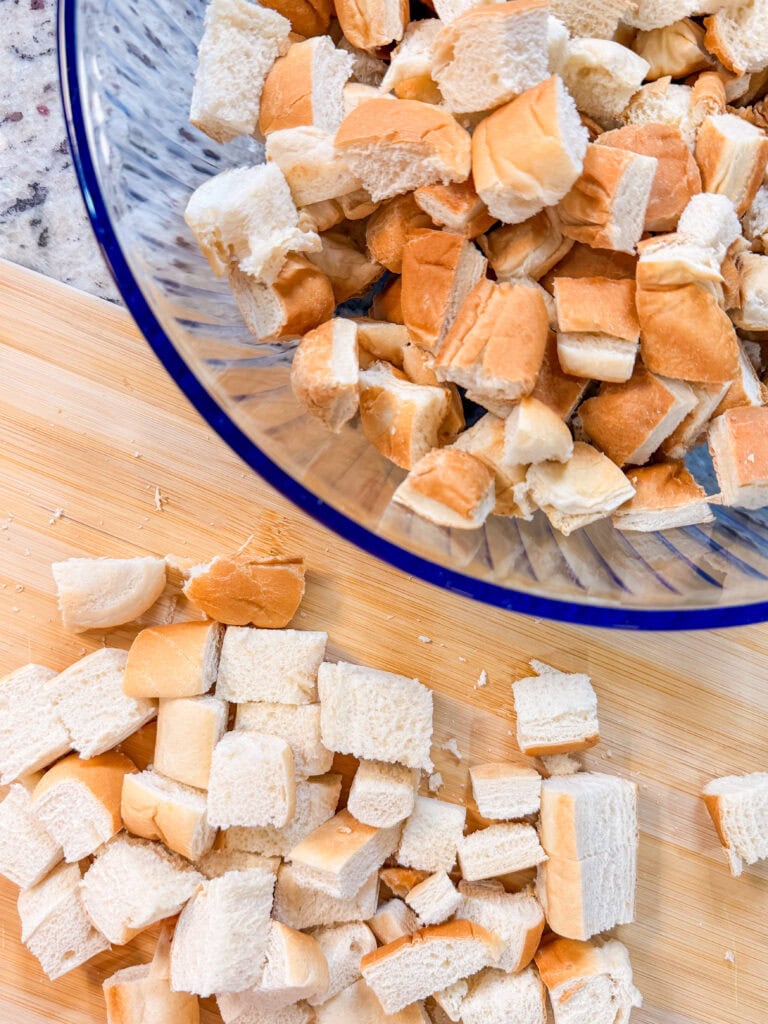 Cubed pieces of bread on a wooden cutting board with more bread cubes in a glass bowl nearby, ready for cooking or baking.