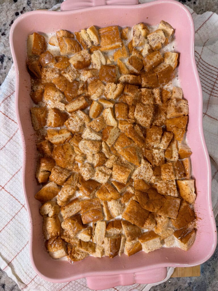 A pink baking dish filled with cubed bread soaked in liquid and sprinkled with cinnamon, sitting on a white and red kitchen towel.