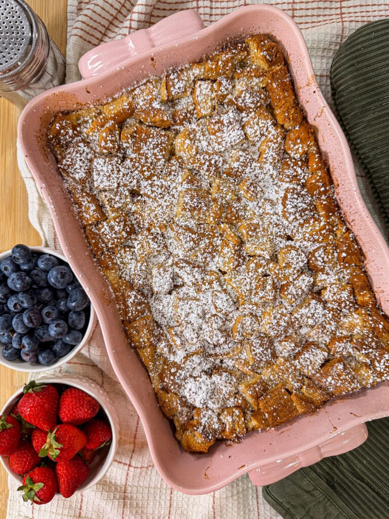 A pink baking dish of baked French toast dusted with powdered sugar sits next to bowls of fresh blueberries and strawberries on a towel with a green oven mitt nearby.