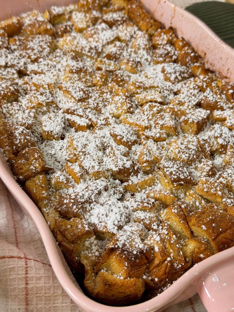 Golden-brown baked bread pudding in a pink dish, dusted with powdered sugar, sits on a textured towel.