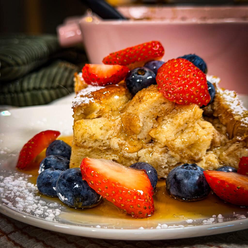 A slice of French toast casserole topped with blueberries, sliced strawberries, powdered sugar, and syrup on a white plate, with a pink baking dish in the background.