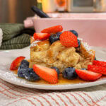 A slice of French toast casserole topped with sliced strawberries, blueberries, powdered sugar, and syrup on a white plate, set on a striped towel.