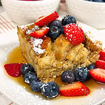 A slice of French toast casserole topped with powdered sugar, strawberries, blueberries, and syrup sits on a white plate. Bowls of strawberries and blueberries are in the background.
