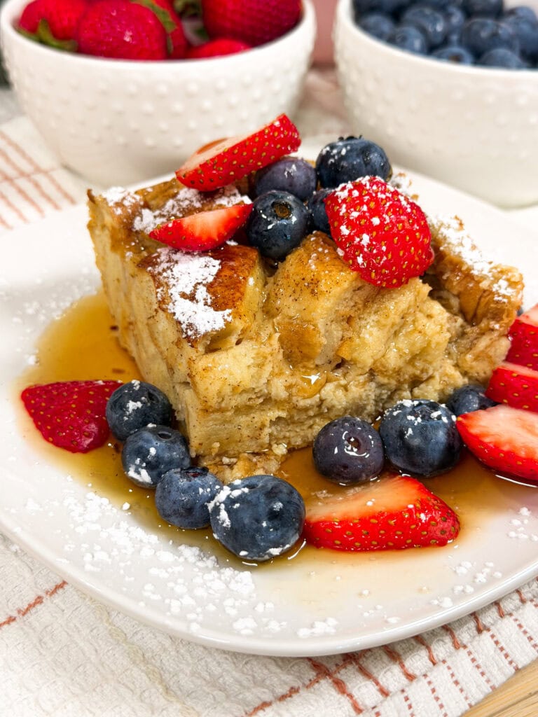 A slice of French toast casserole topped with powdered sugar, strawberries, blueberries, and syrup sits on a white plate. Bowls of strawberries and blueberries are in the background.