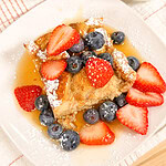 A square slice of French toast topped with strawberries, blueberries, powdered sugar, and syrup sits on a white plate. Bowls of strawberries and blueberries are in the background.