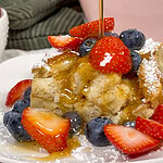 A slice of French toast casserole topped with fresh strawberries, blueberries, powdered sugar, and syrup being poured over it, served on a white plate.