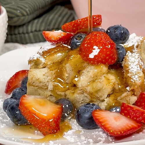 A slice of French toast casserole topped with fresh strawberries, blueberries, powdered sugar, and syrup being poured over it, served on a white plate.