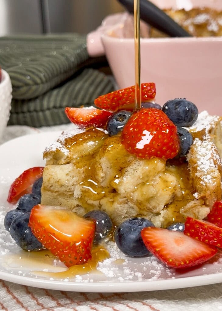 A slice of French toast casserole topped with fresh strawberries, blueberries, powdered sugar, and syrup being poured over it, served on a white plate.