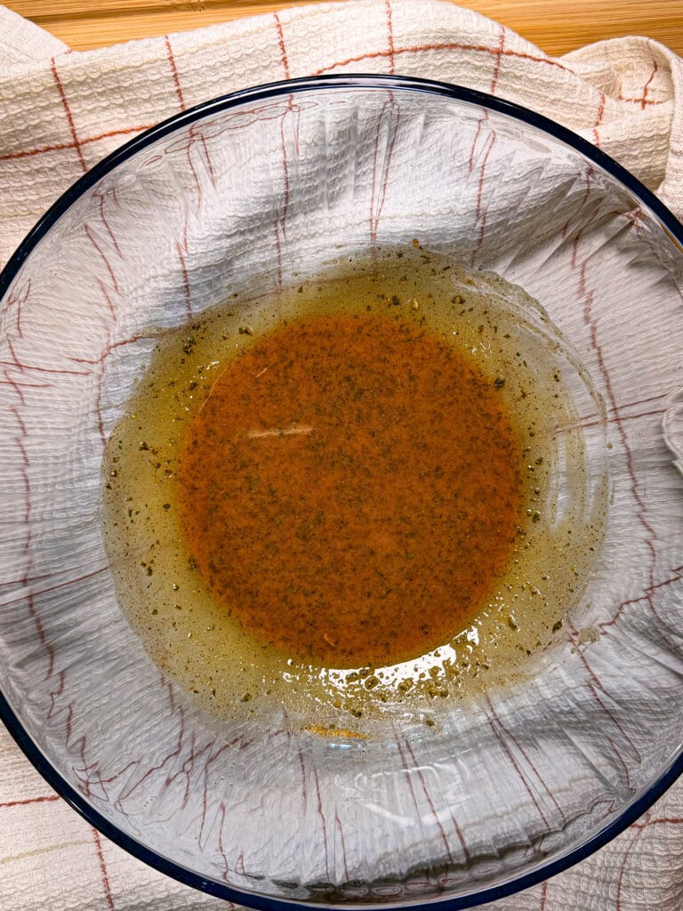 A clear bowl with seasoned oil or marinade sits on a white and red checkered cloth. The liquid appears golden-brown with visible herbs and spices mixed in.